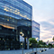 A tall tree-lined office building of blue-toned mirrored glass reflecting a pale orange and blue sunset