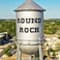 A large steel water tower with the words “Round Rock” on the side, with a sprawling tree-filled town in the background beneath a clear bright blue sky