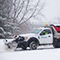 A white snowplow driving down a tree-lined road during a heavy snowstorm