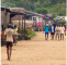 Groups of people walk down a village road lined with wooden houses at the base of a lush mountain