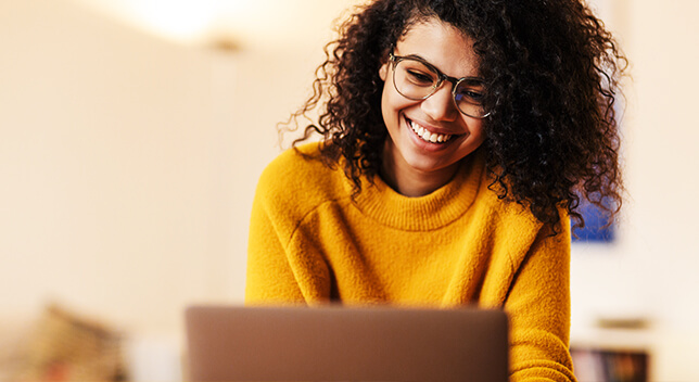 Giovane sorridente con occhiali e un maglione giallo mentre guarda il monitor di un laptop