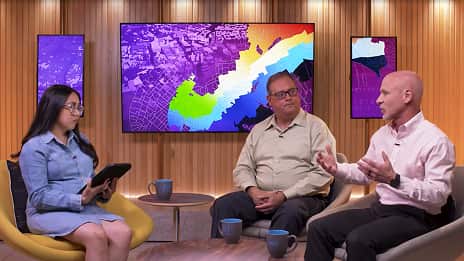 A discussion panel features three individuals seated in a studio setting, with a vibrant background of colorful graphical displays