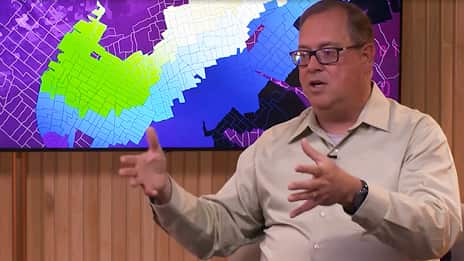 A man with glasses, wearing a light green shirt, sits in front of a computer screen showing a vibrant, detailed map