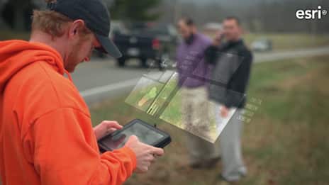 A man in an orange hoodie examines a tablet displaying maps and data, while two colleagues stand nearby, with a road visible in the background