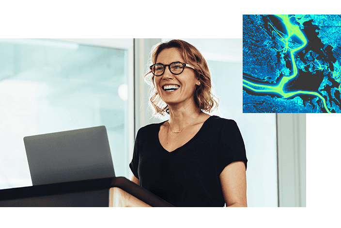 A woman wearing glasses stands at a podium, smiling, with a laptop displaying a blue-green satellite image and a road map beside her
