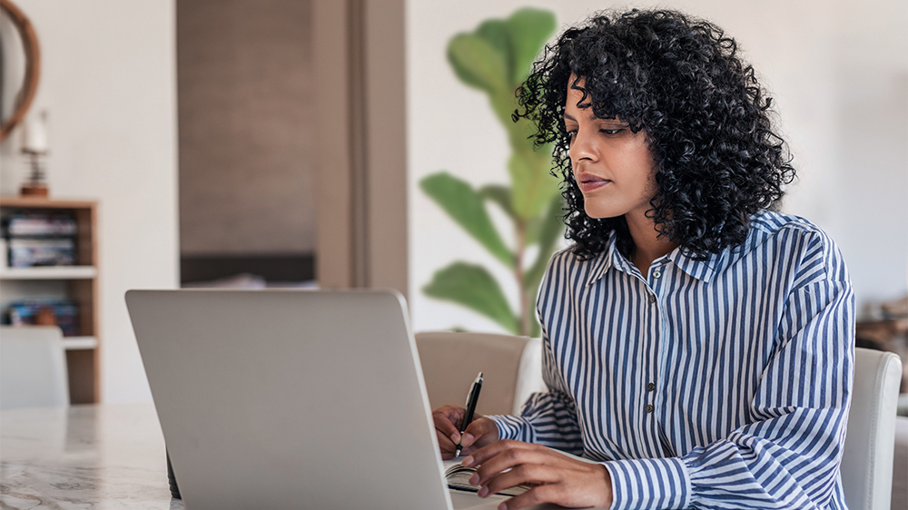 A businessperson sitting at her desk with a laptop writing on a notepad