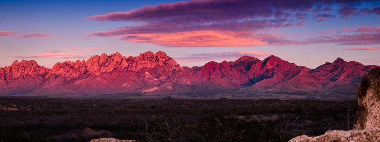 A desert landscape at sunset, showcasing a range of reddish-brown mountains silhouetted against a soft, pinkish-purple sky.