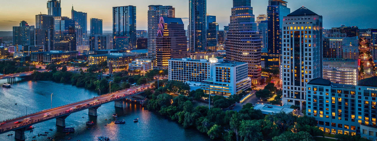 Nighttime cityscape of Austin, Texas, showing the city skyline along a river with bridges.