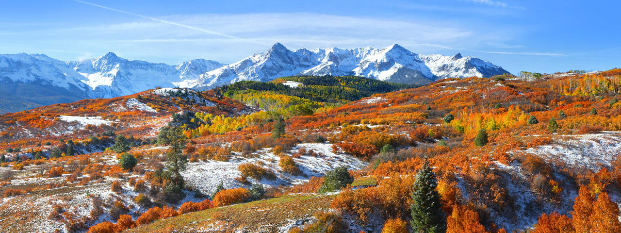 Panoramic view of a snow-capped mountain range in autumn, with vibrant orange, red, and yellow foliage covering the hillsides.